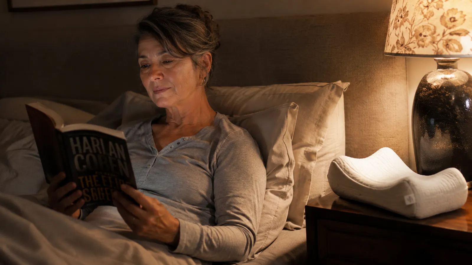 Woman reading in bed with a knee pillow visible beside her on the nightstand, warm lamp light, cozy bedroom setting
