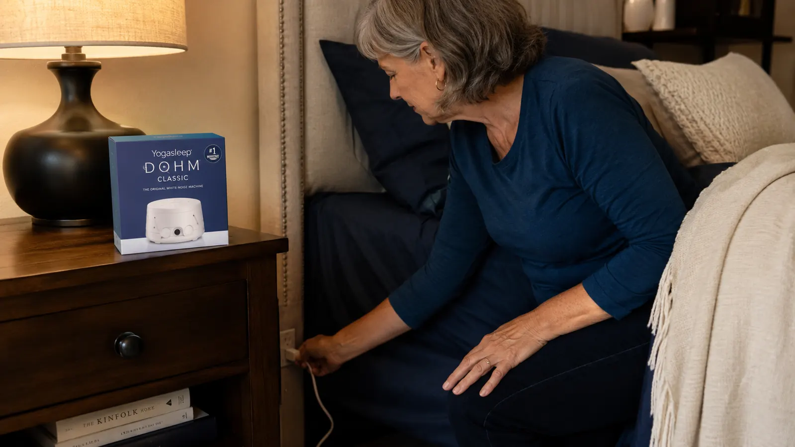Person plugging the Yogasleep Dohm Classic into a wall outlet behind a nightstand, showing the short power cord reaching the outlet